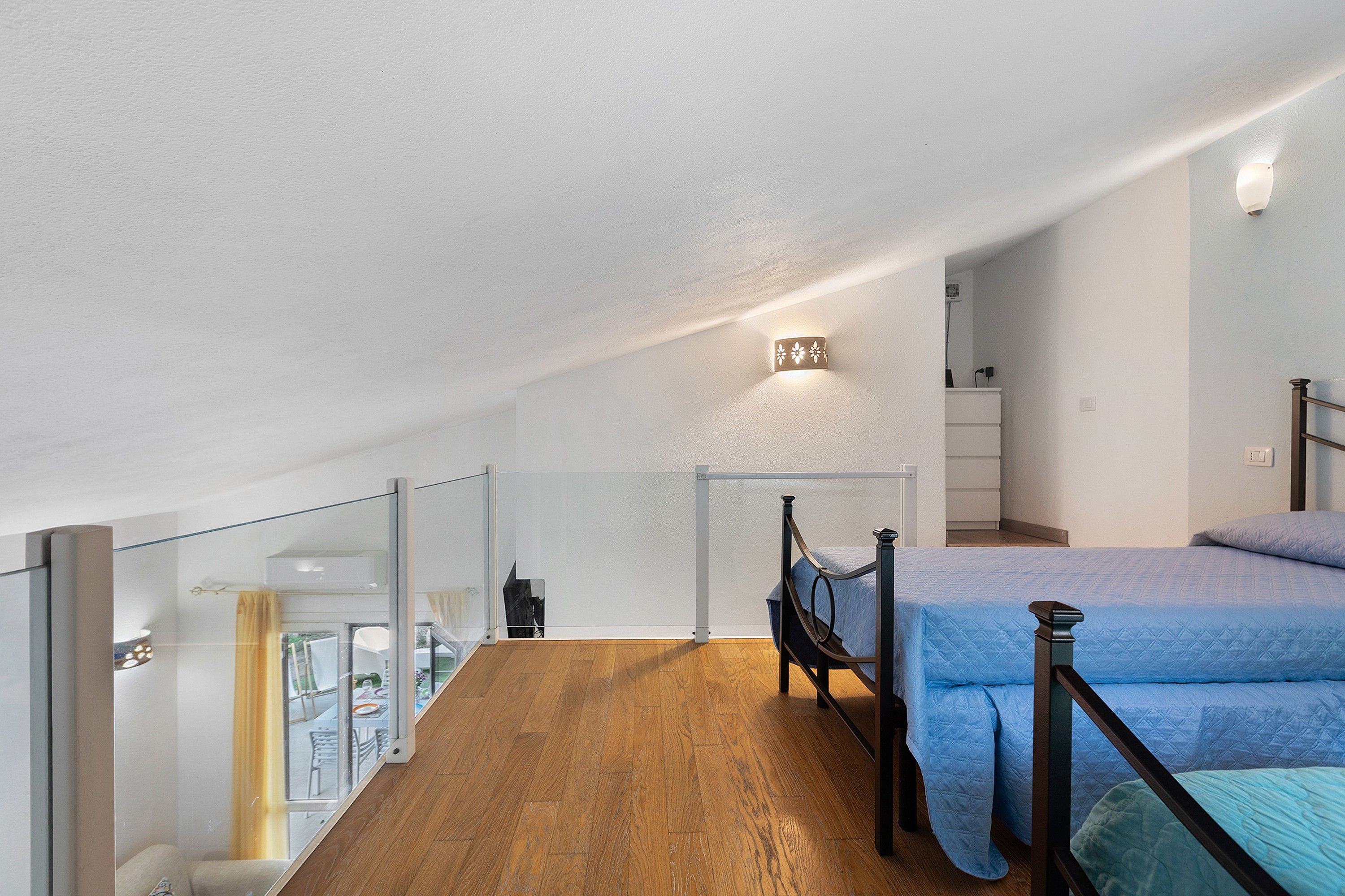 A bedroom with a wooden floor, a bed with a blue bedspread, a nightstand, and a wall-mounted light fixture.