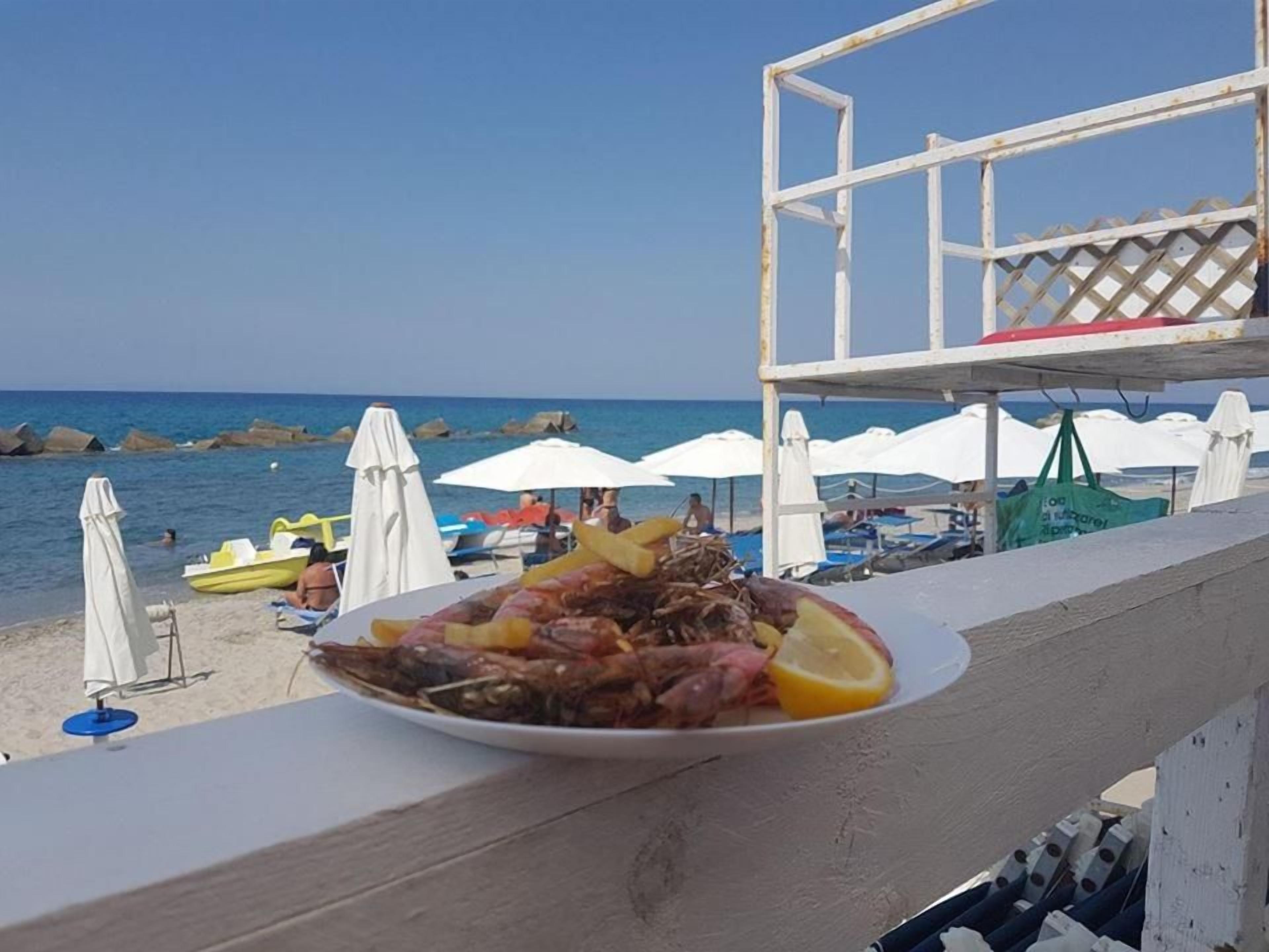 Una mesa de comedor frente a la playa, con un plato de comida, sombrillas y vista al mar.