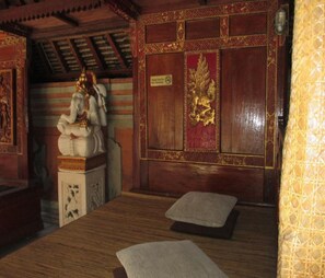 Una habitación con techo de madera, una estatua, un panel decorativo rojo y dorado, y una cama con un almohadón.