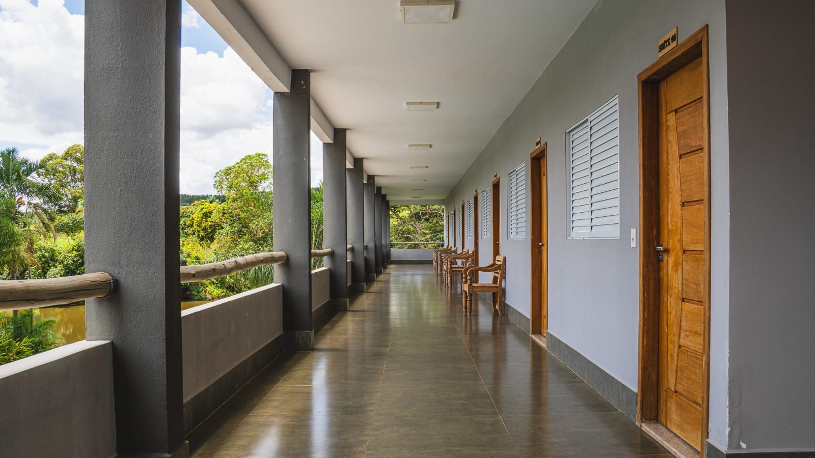 A corridor with a wooden railing, doors, and a view of greenery.
