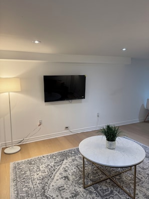Ein modernes Wohnzimmer mit einem an der Wand befestigten Flachbildfernseher, einem runden weißen Tisch mit einer Pflanze, einem gemusterten Teppich und einer Stehlampe.