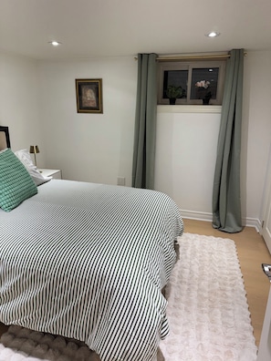 Ein Schlafzimmer mit Bett, Nachttisch, Lampe, einem gerahmten Bild an der Wand und einem Fenster mit Vorhängen.