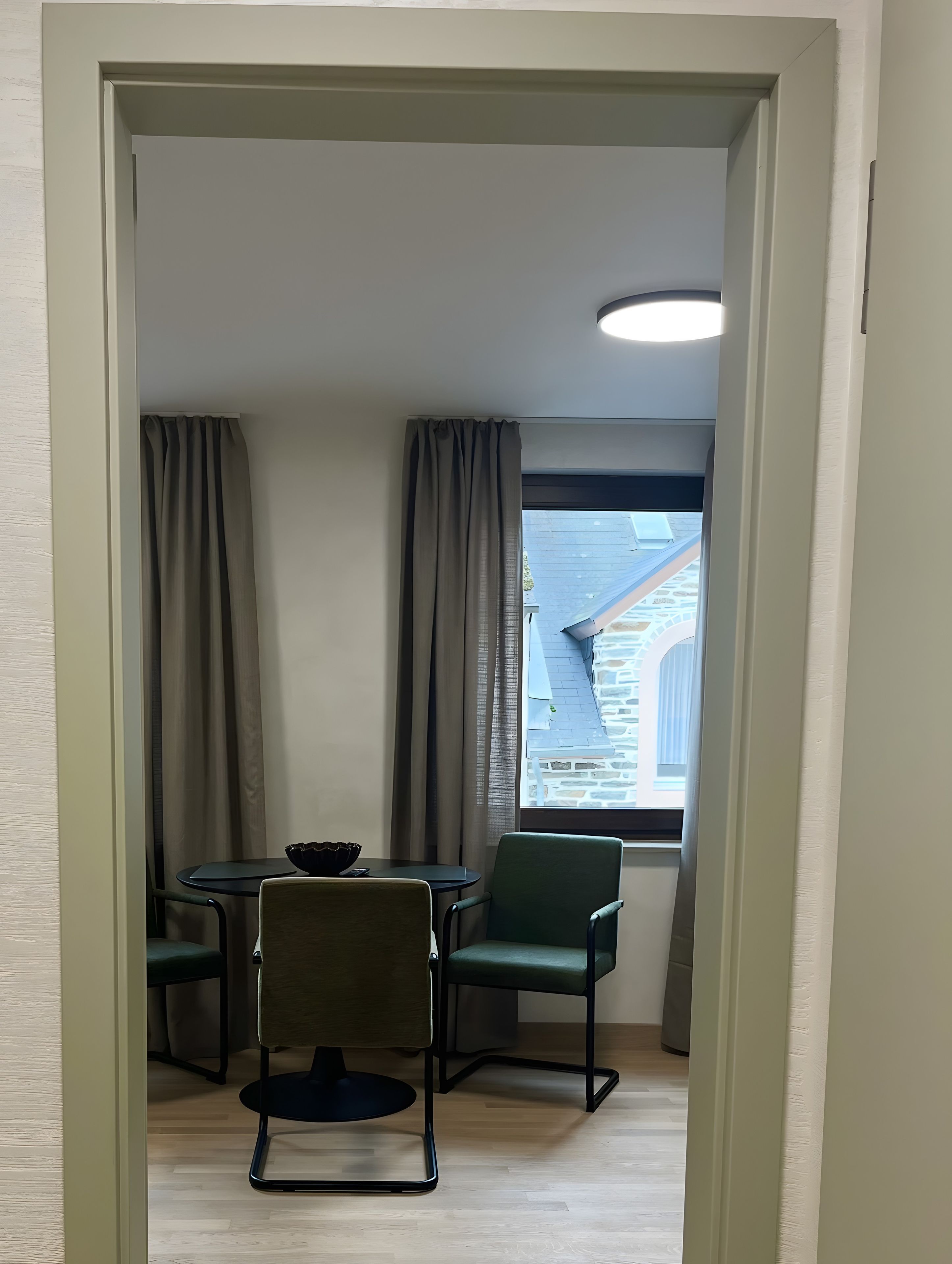 A room with a dining table and chairs, a window with a view of a building, and a ceiling light.