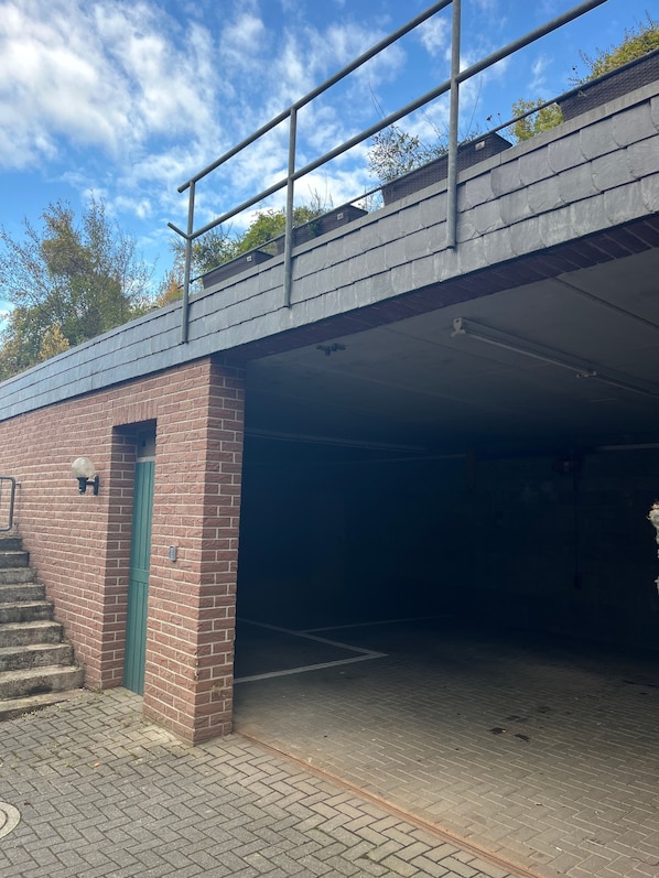 Un espace de stationnement couvert, avec un mur en briques, une porte verte et une rambarde métallique sur un pont.