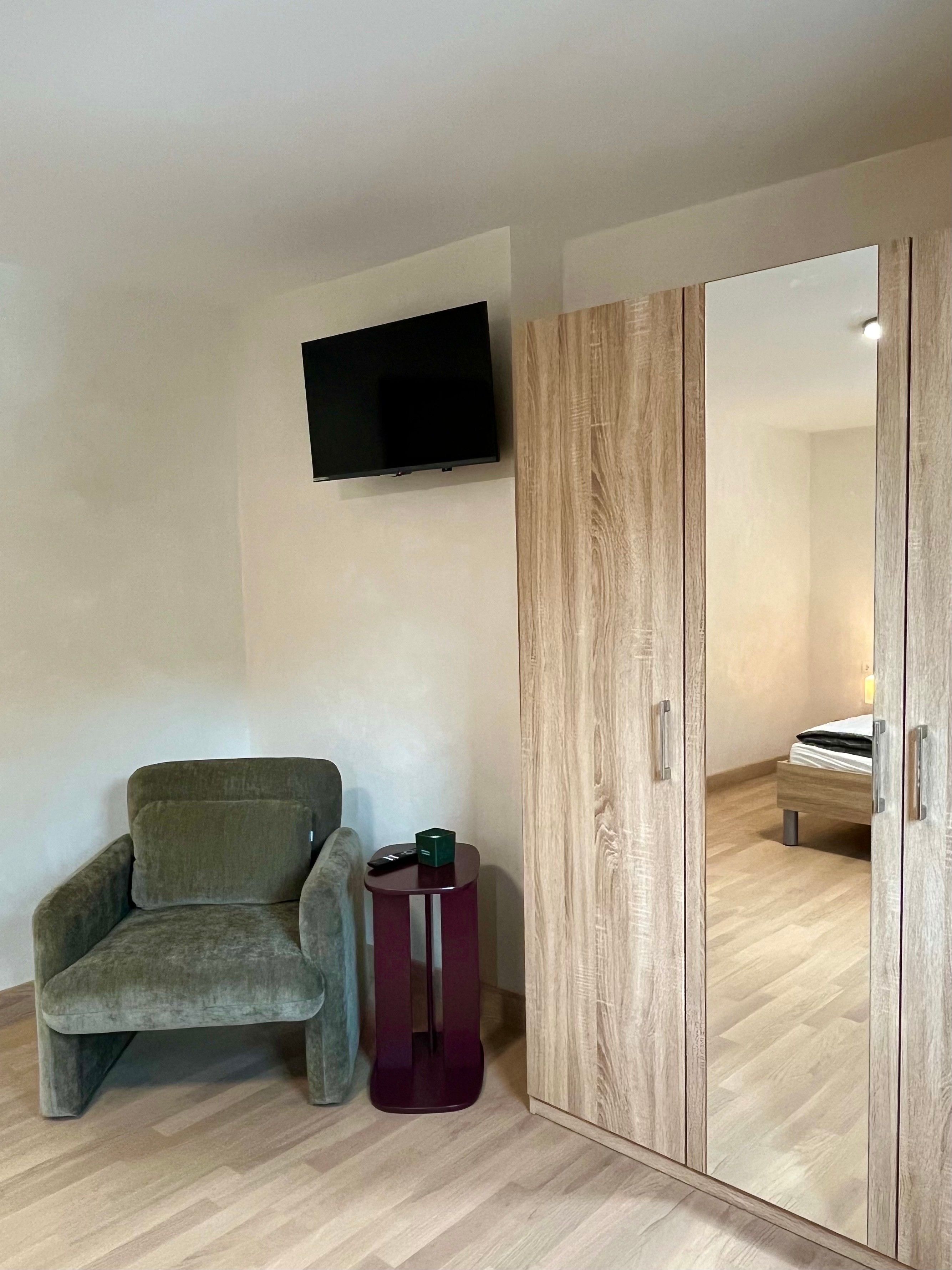 Ein Zimmer mit grauem Sessel, einem kleinen Beistelltisch mit Fernbedienung, einem Holzschrank und einem flachbildfernseher an der Wand.