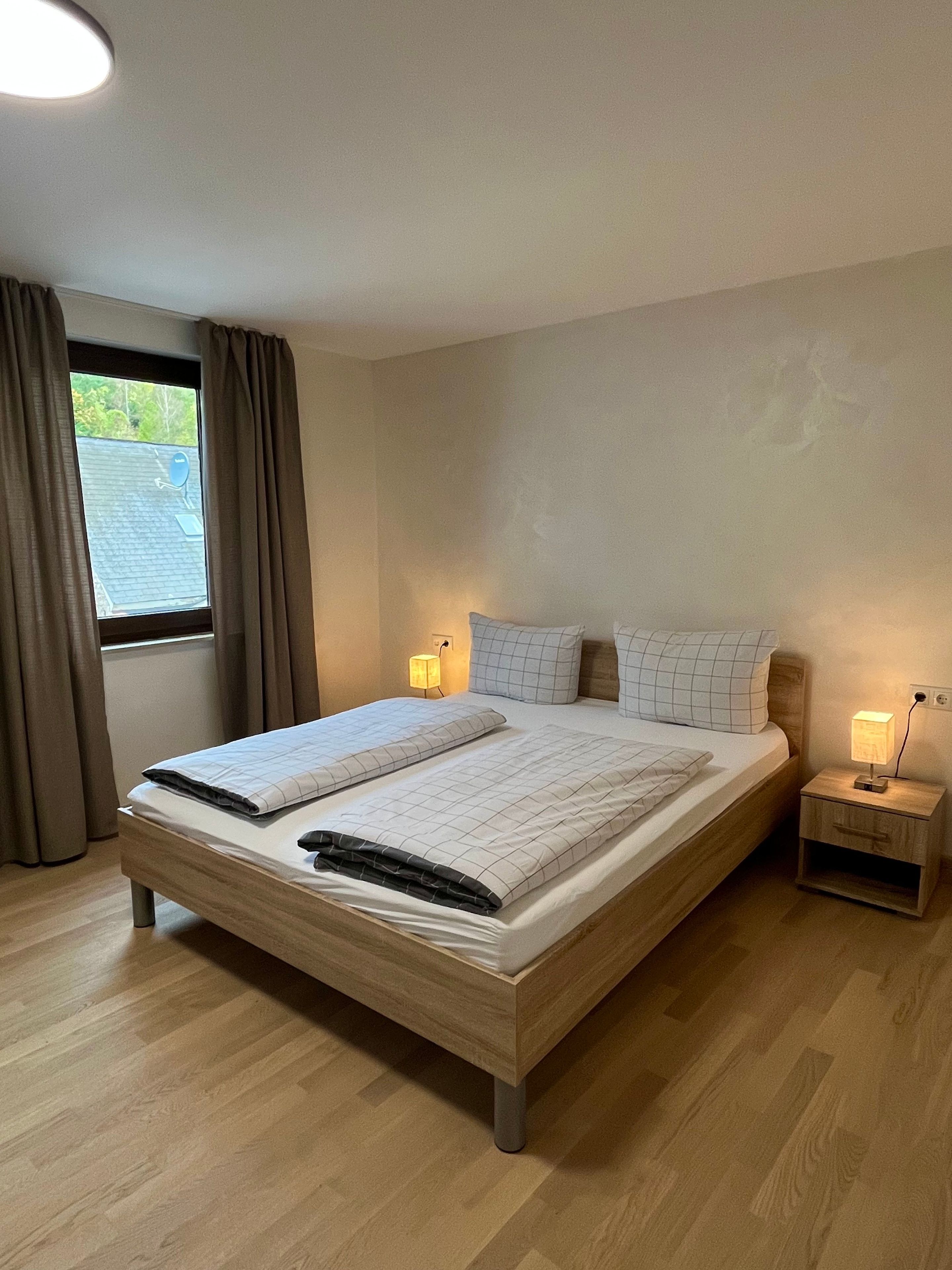 Ein Schlafzimmer mit einem hölzernen Bett, zwei Nachttischen mit Lampen, einem Fenster mit Vorhängen und Blick ins Freie.