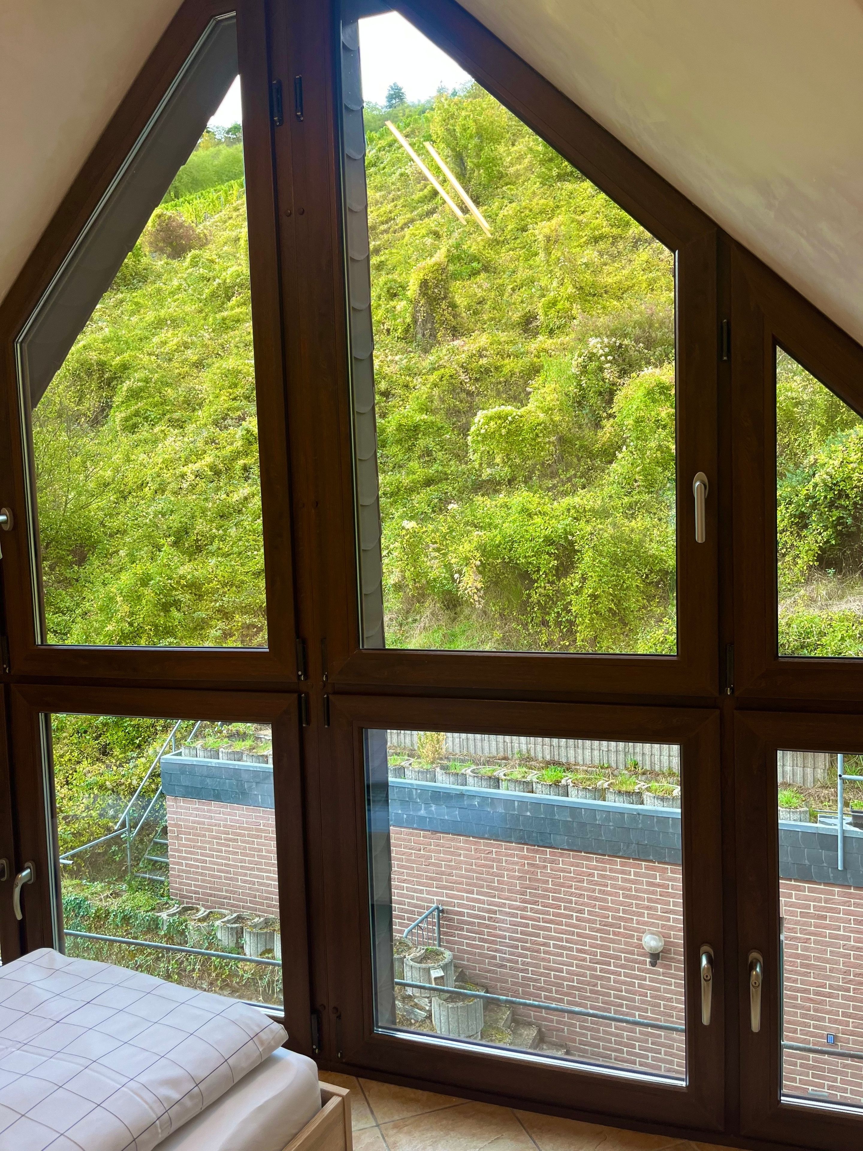 Ein Zimmer mit großen Fenstern, einem Bett und Blick auf eine Ziegelwand und Grünflächen.