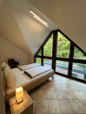 Ein Schlafzimmer mit schrägen Wänden, einem Bett mit Kopfteil, einem Nachttisch mit Lampe und großen Fenstern, die einen Blick auf Grünflächen bieten.