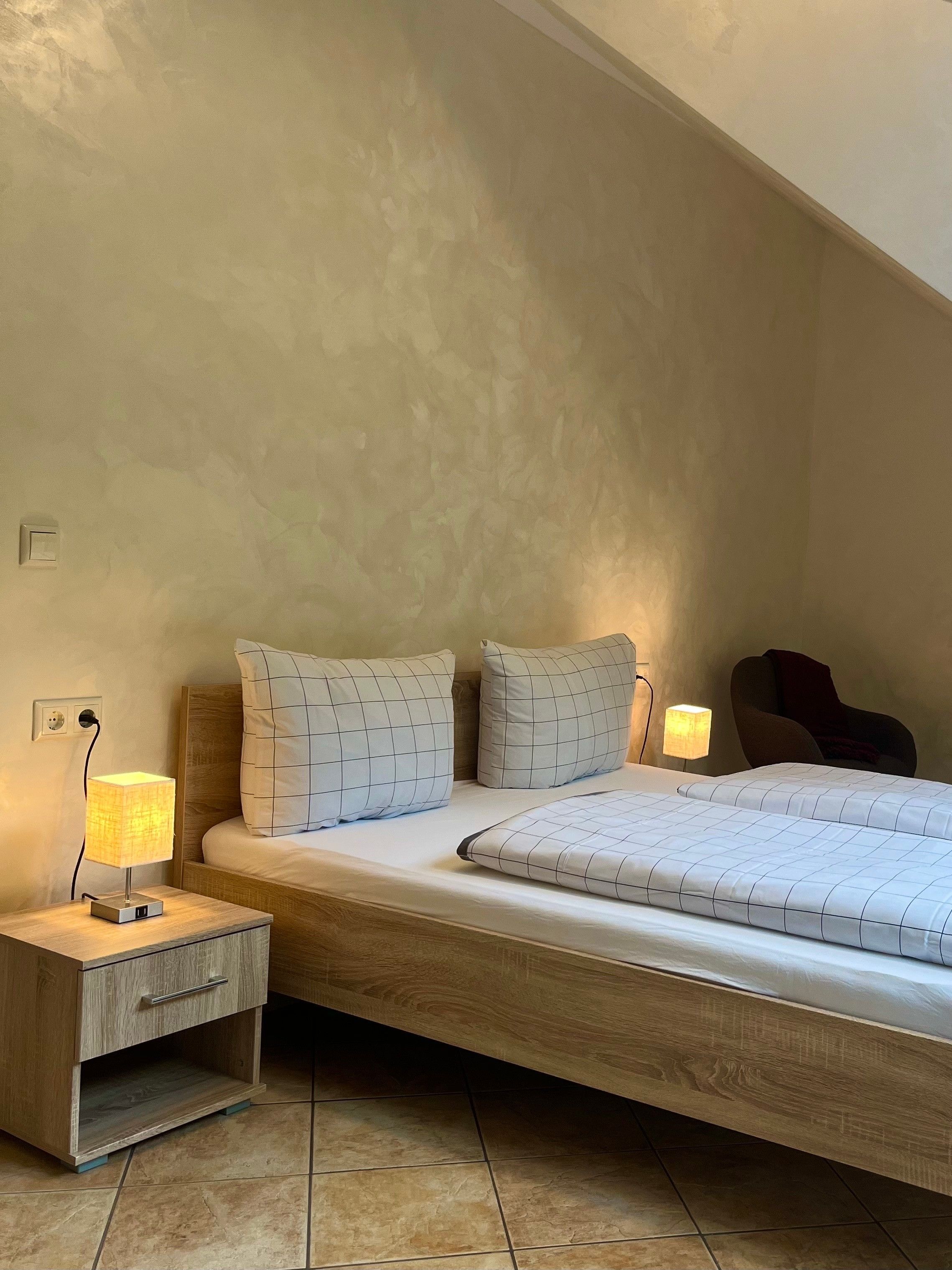 Ein Schlafzimmer mit schrägem Dach, einem hölzernen Bettgestell, einem Nachttisch mit Lampe und einem Sessel.