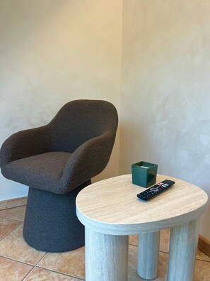Ein Zimmer mit einem grau gepolsterten Sessel, einem kleinen runden Holztisch und einer Fernbedienung auf dem Tisch.