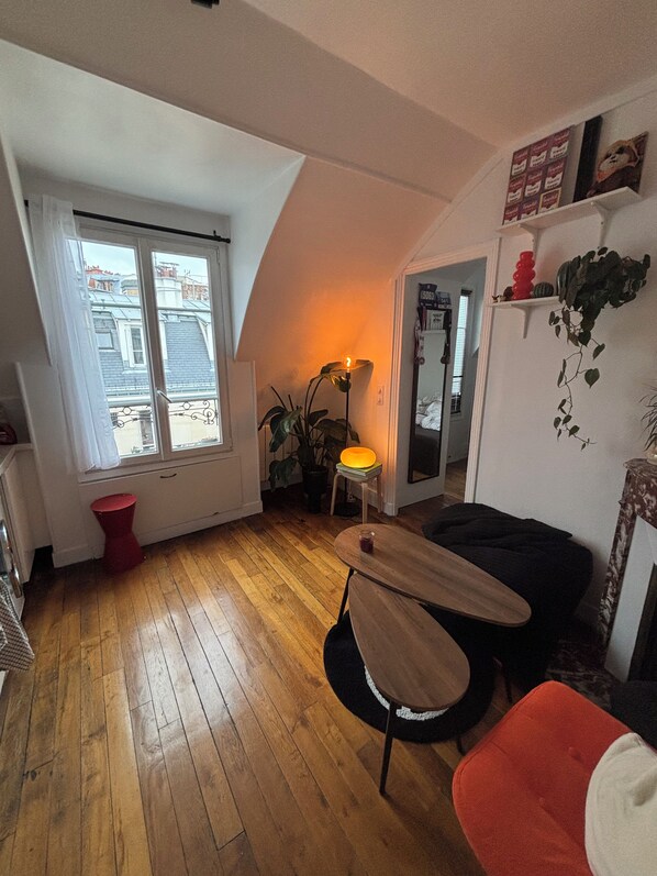 Un espacio de sala acogedor con piso de madera, un taburete rojo, una mesa con lámpara y un sofá negro.