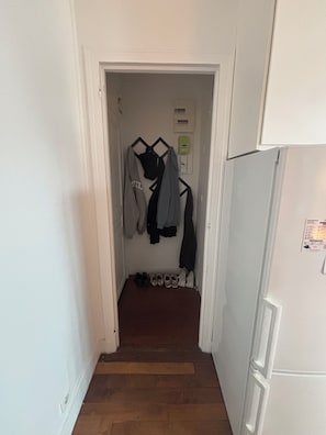 Un couloir étroit, doté d’un plancher en bois, mène à une pièce dont la porte est blanche. Des chaussures sont alignées le long du mur, et un porte-manteau est garni de différents vestons.