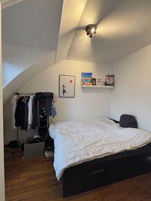 Un dormitorio con cama, un armario con ropa colgada, una estantería con libros y una lámpara en el techo.
