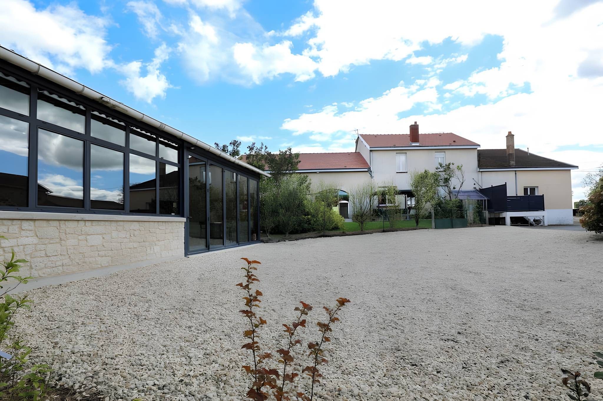 Un bâtiment avec une allée en gravier, une structure en verre et une maison d’habitation en arrière-plan.