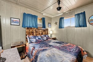 Un dormitorio con cabecera de madera, cama con colcha de motivos forestales, mesita de noche, lámpara y ventana con cortinas.