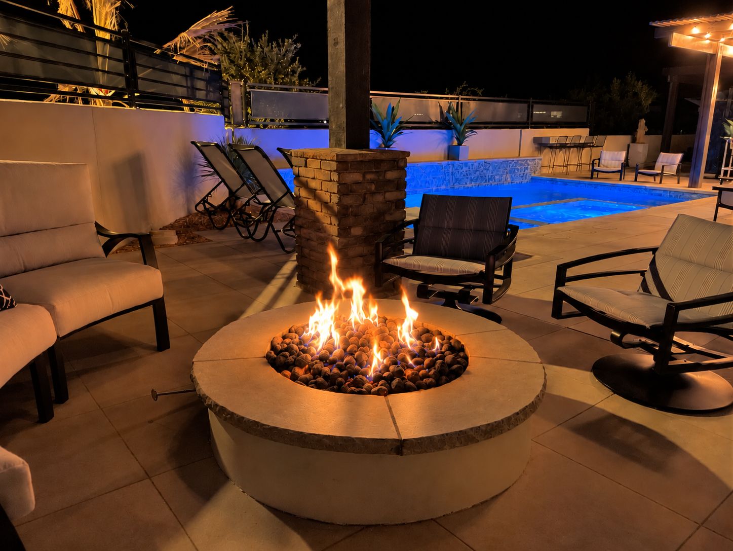 A circular fire pit with logs, surrounded by lounge chairs and a pool in the background.