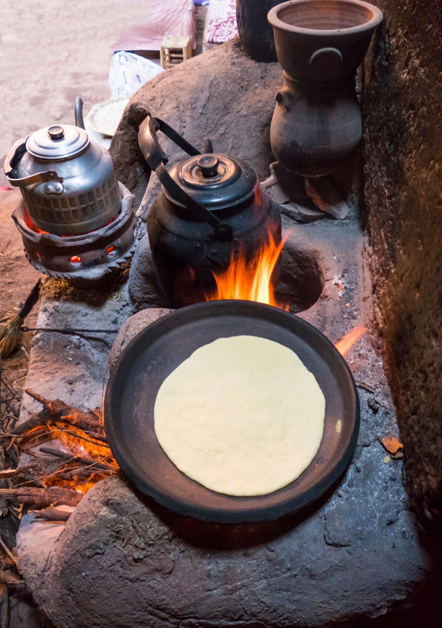 Un montaje de cocina tradicional con un pan plano en una sartén, un hervidor y una olla sobre fuego abierto.