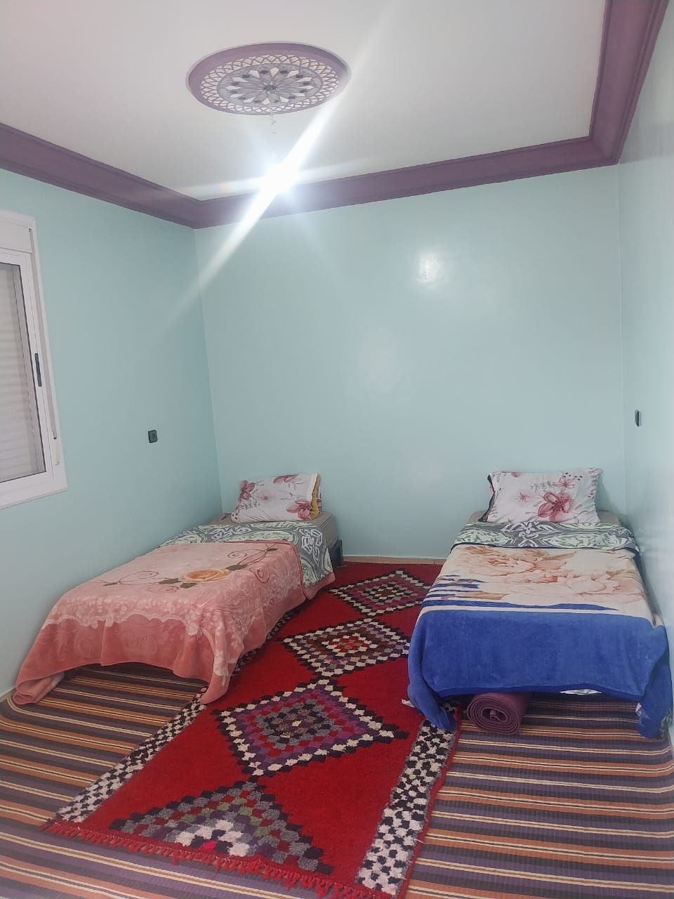 Quarto com duas camas de solteiro, carpete estampado, luminária no teto e janela com persianas.
