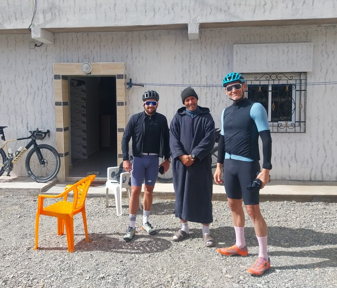 Trois personnes se tiennent devant un bâtiment, avec un vélo et une chaise à proximité.