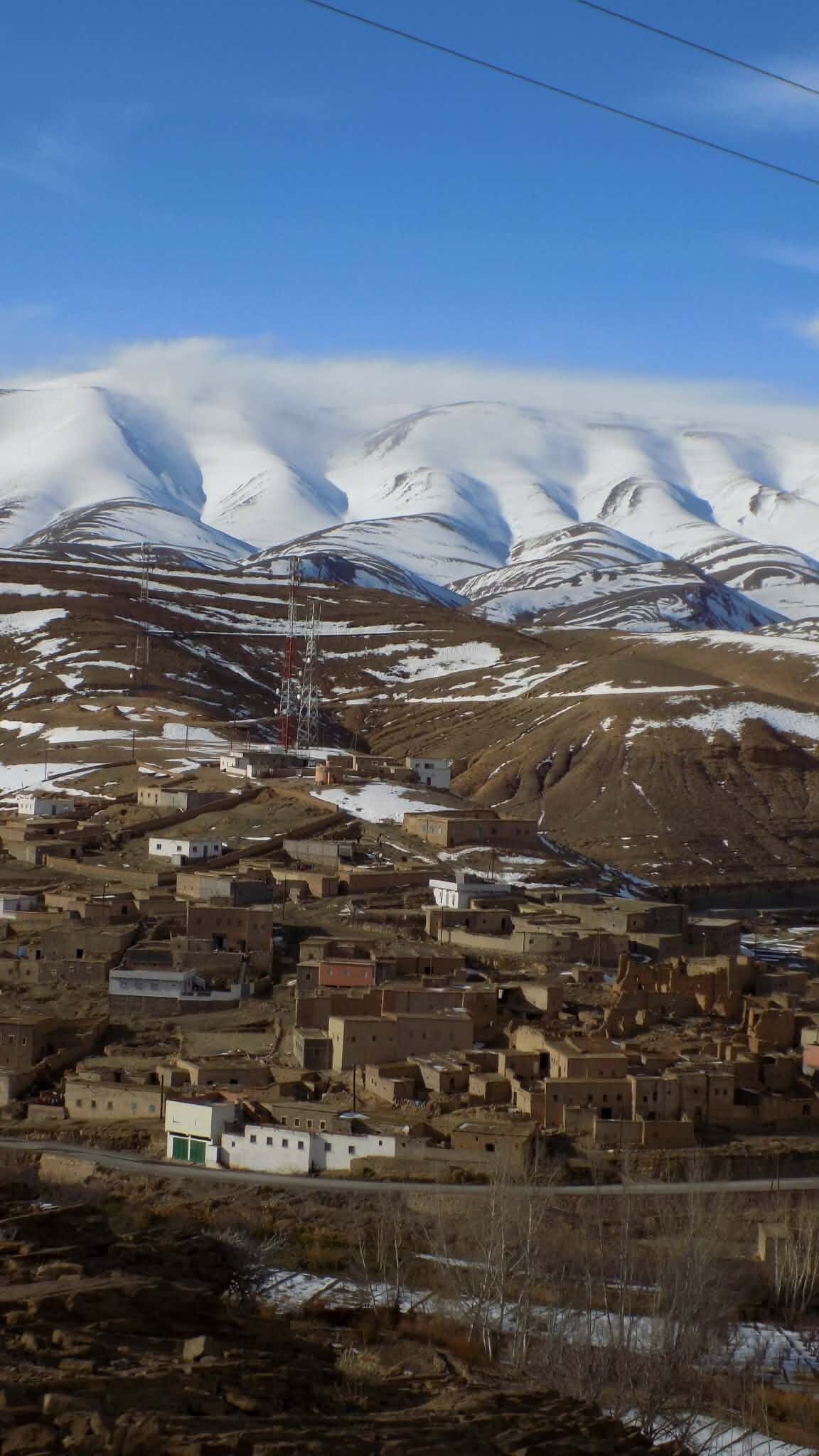 Un pueblo montañoso con picos nevados al fondo, algunos edificios y un cielo despejado.