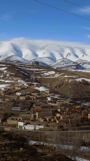 A mountainous village with snow-capped peaks in the background, a few buildings, and a clear sky.
