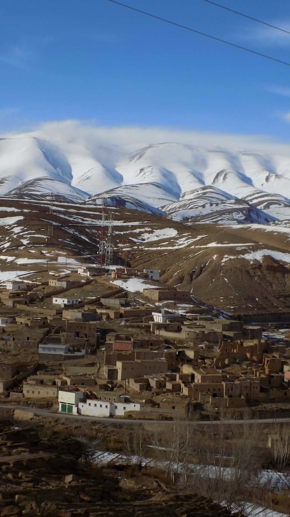 A mountainous village with snow-capped peaks in the background, a few buildings, and a clear sky.