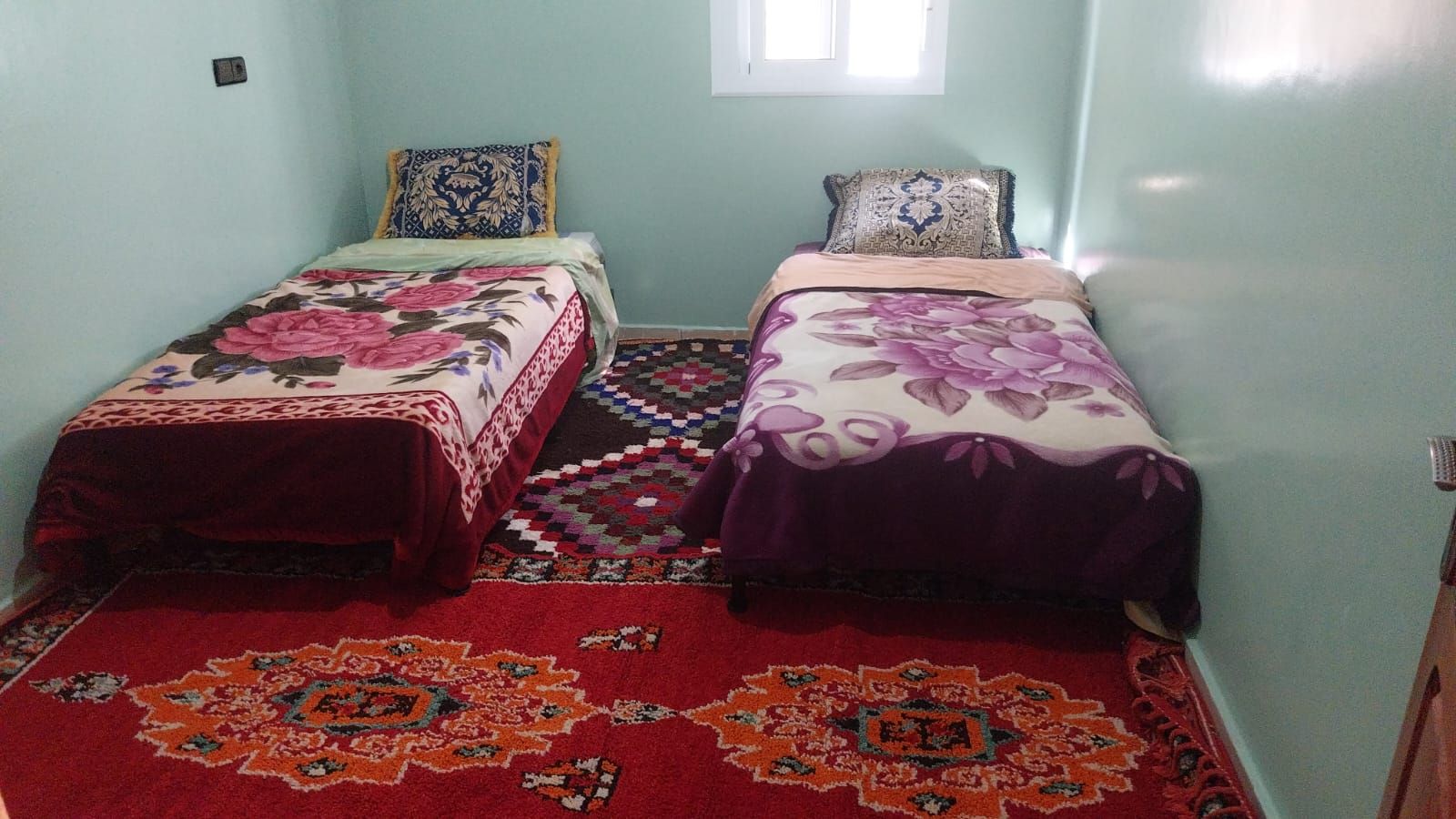 A room with two beds, each with a floral bedspread, a patterned rug, and a window with daylight.