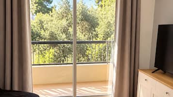 Ein Zimmer mit einem Bett, einem Fernseher auf einer Holzkommode und einer Glastür zu einem Balkon mit Blick auf Bäume.