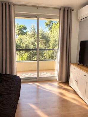 Ein Zimmer mit einem Bett, einem Fernseher auf einer Holzkommode und einer Glastür zu einem Balkon mit Blick auf Bäume.