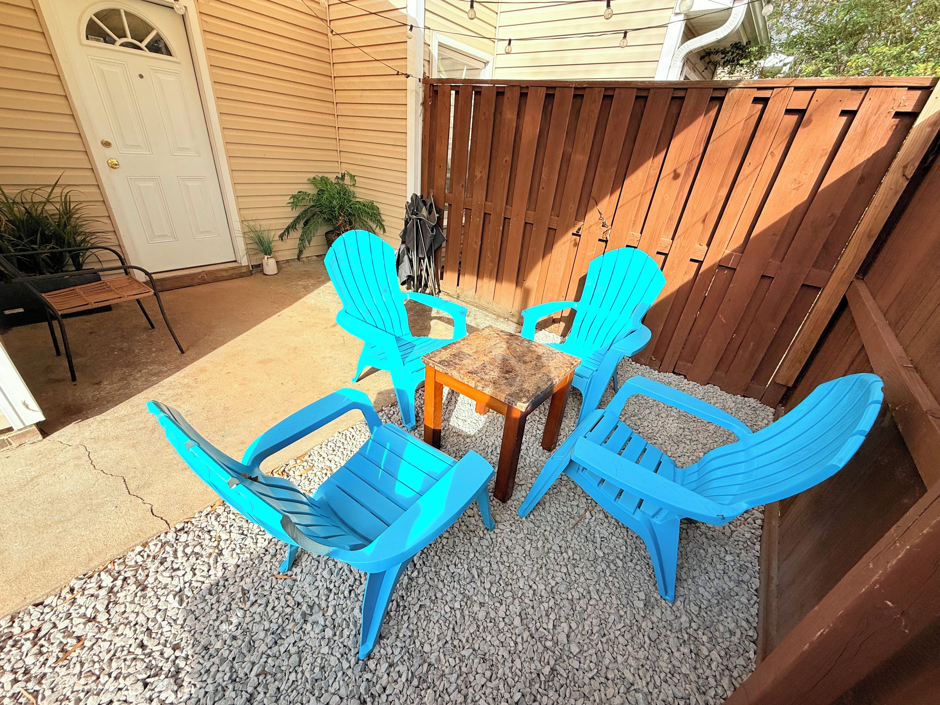 Une terrasse avec une table en bois et des chaises en plastique bleues, entourée d’une clôture en bois et d’une porte blanche.
