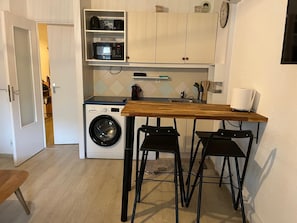 A compact kitchen with a washing machine, a wooden table, and two bar stools. - Apartment 'Valescure 2 Pièces Saint Raphael' with Wi-Fi (Saint-Raphaël)