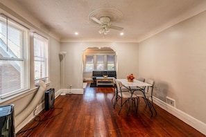 Quarto com piso de madeira, ventilador de teto, mesa de jantar com cadeiras e um sofá.