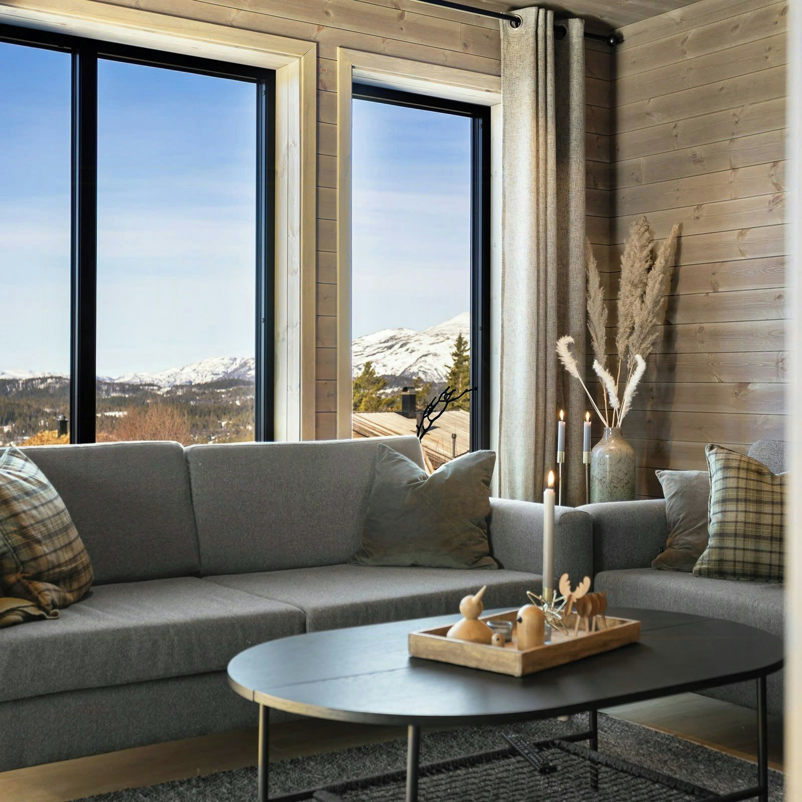 A modern living room with a grey sofa, a round coffee table, and a view of snow-capped mountains through large windows.