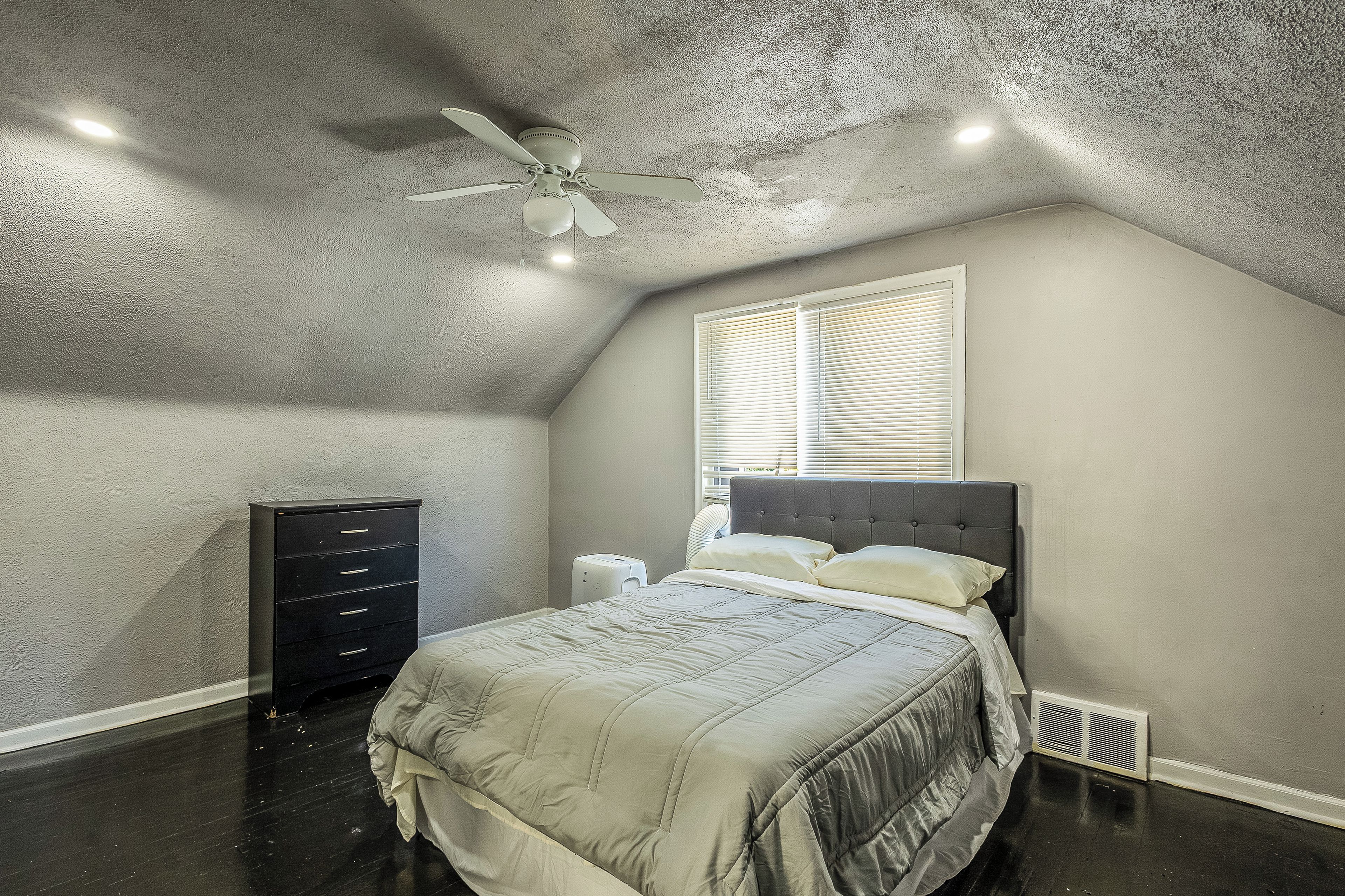 A bedroom with a sloped ceiling, a bed with a headboard, a dresser, and a window with blinds.