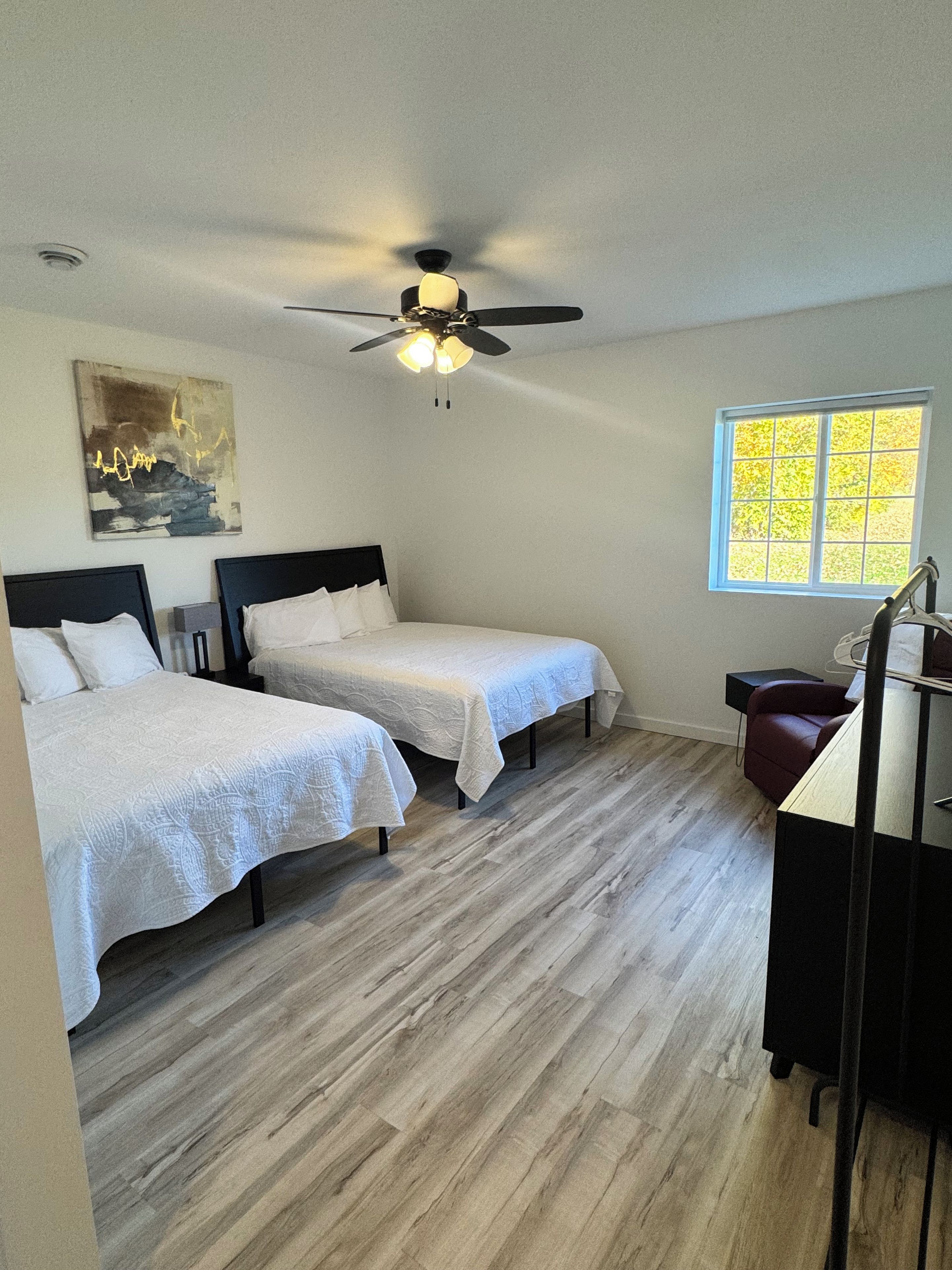 A room with two beds, a ceiling fan, a window with a view, and a desk with a chair.