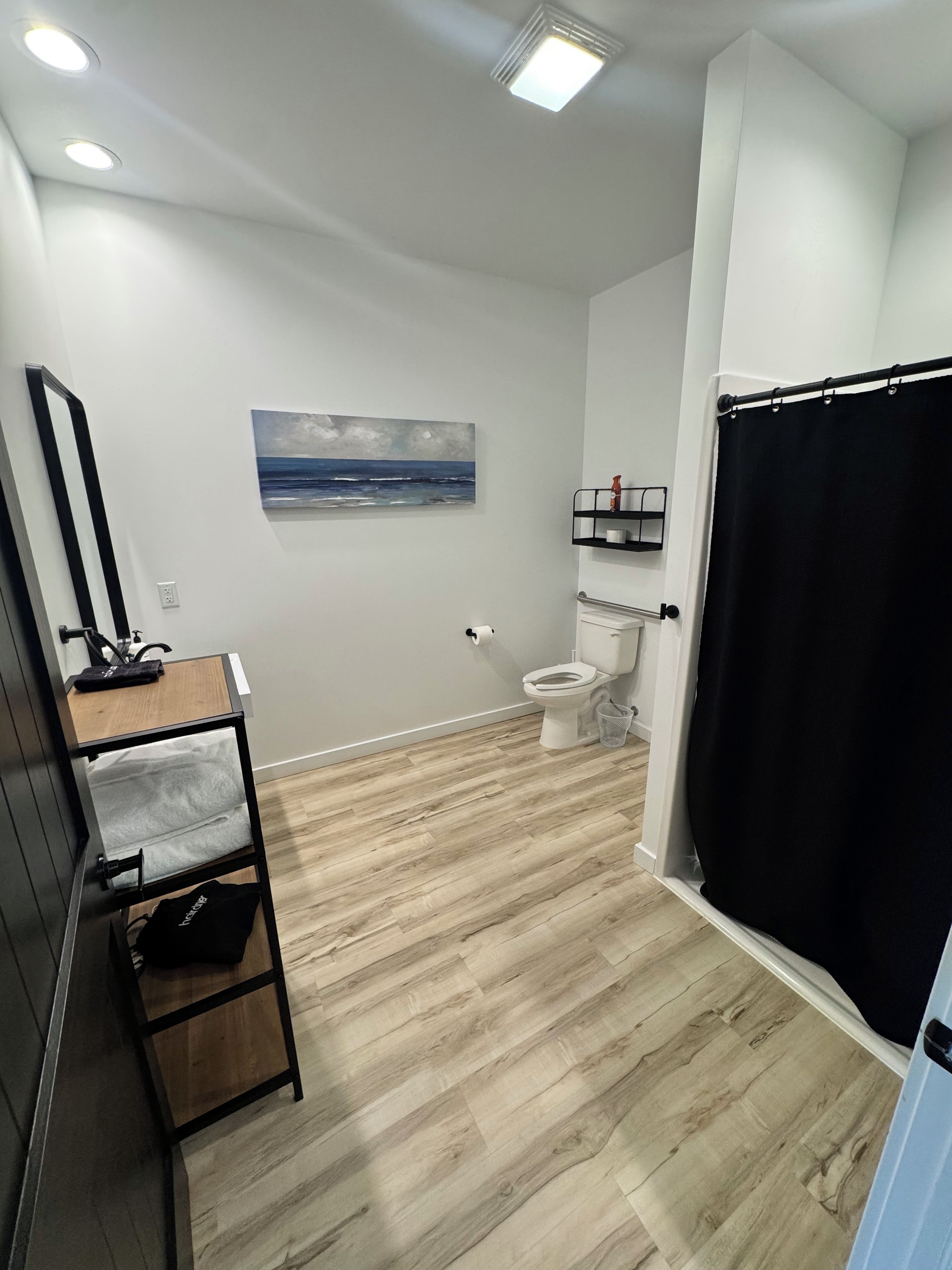 A bathroom with a wooden floor, a toilet, a black shower curtain, a painting on the wall, and a mirror.