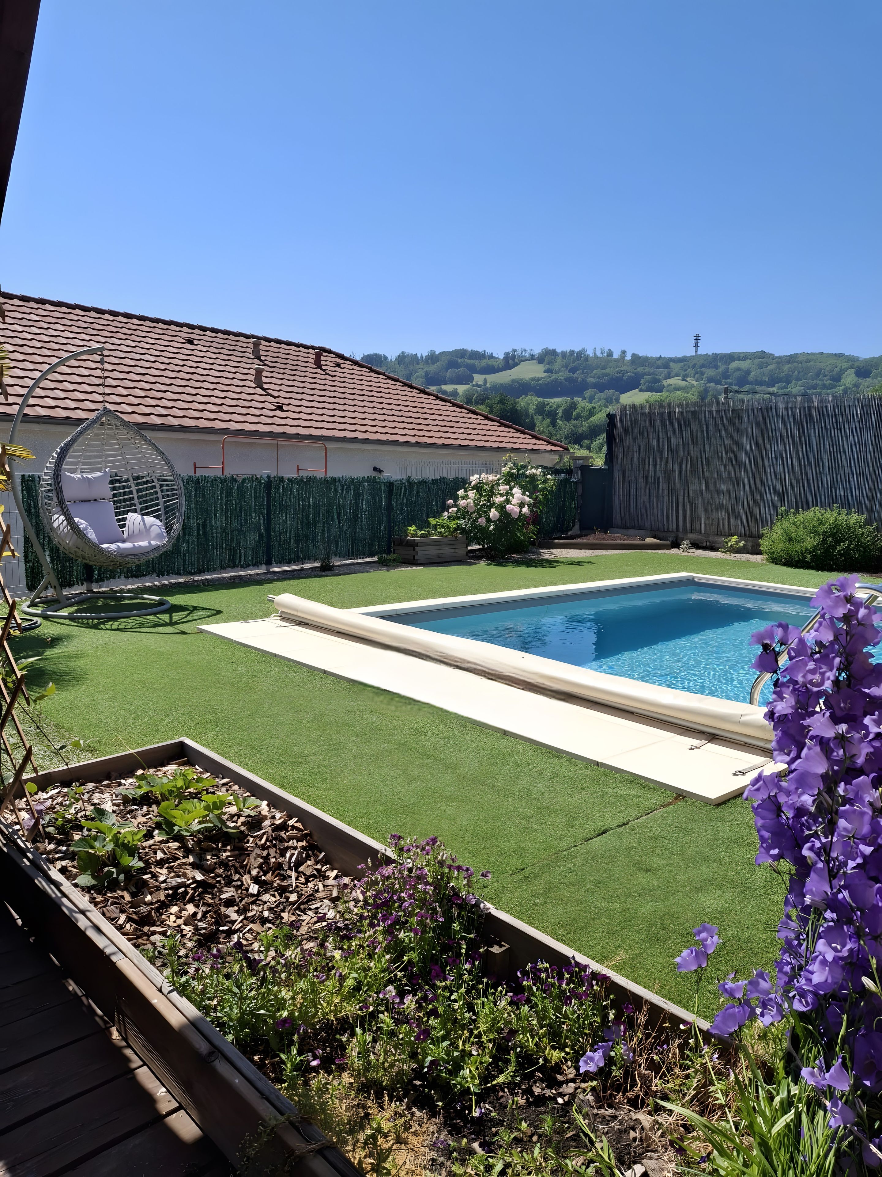 Un patio trasero con piscina, una silla colgante y un jardín lleno de flores.