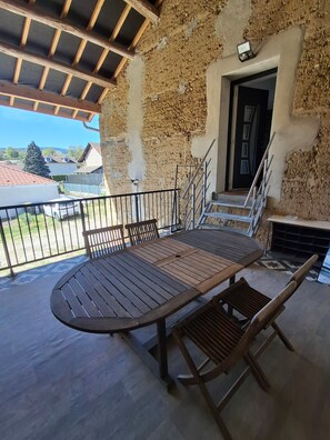 Un área de estar al aire libre con techo, una mesa y sillas de madera, baranda de metal y vista del barrio circundante.