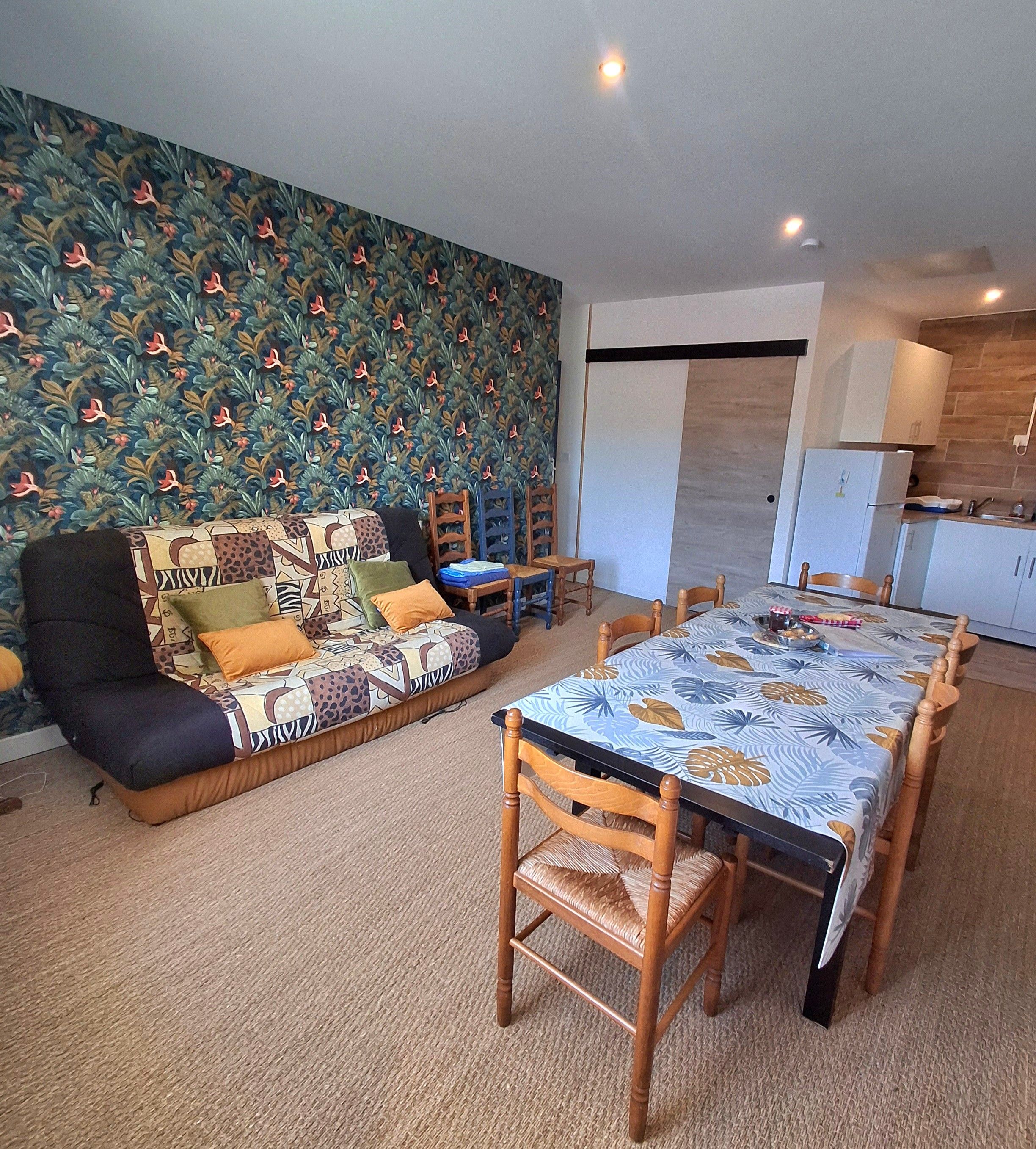 Una sala de estar con un papel tapiz decorativo, un sofá, una mesa de comedor con sillas y una cocina con armarios y un refrigerador.