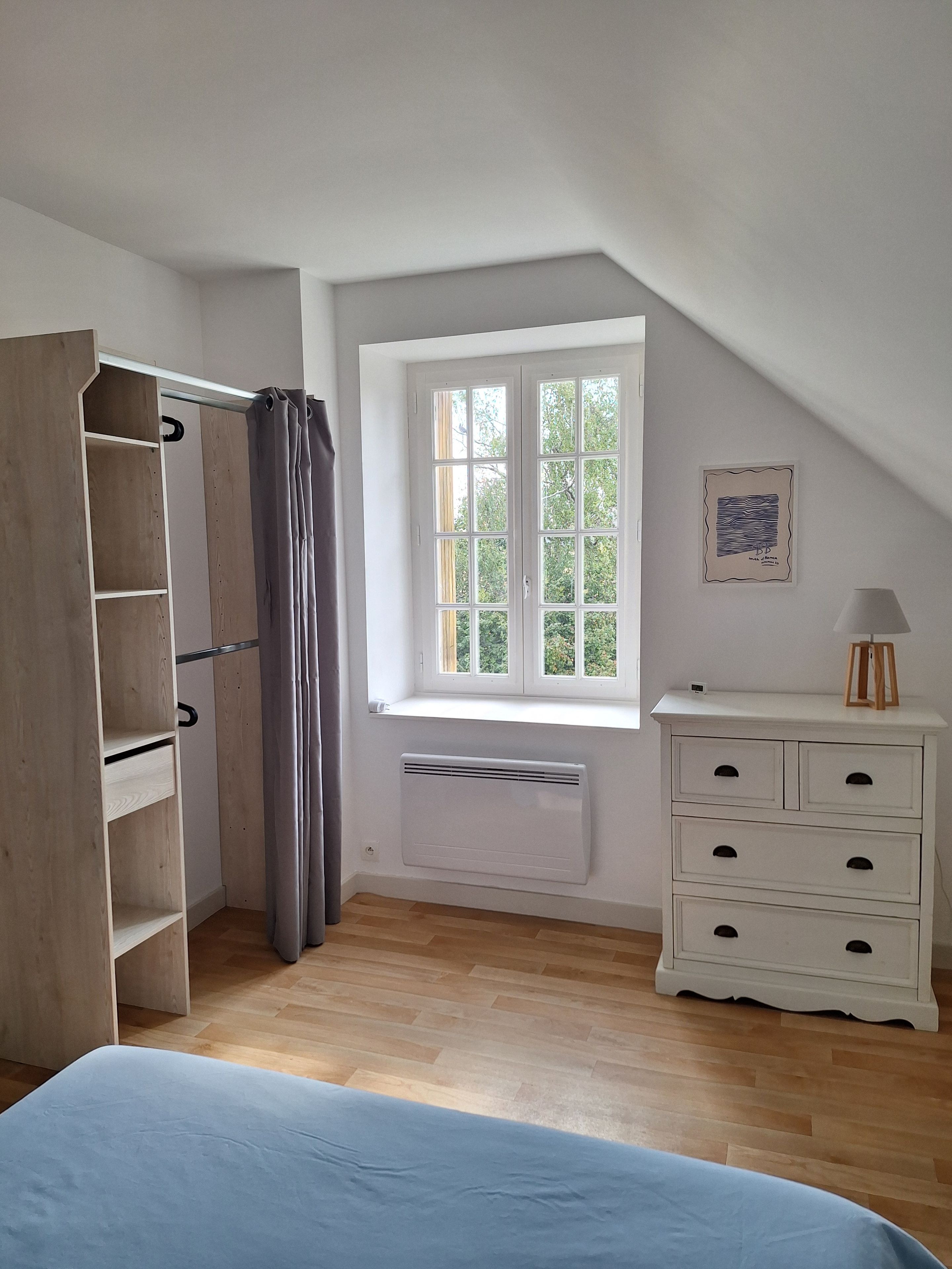 A bedroom with a sloped ceiling, a window with a view of greenery, a white dresser, and a wooden wardrobe.