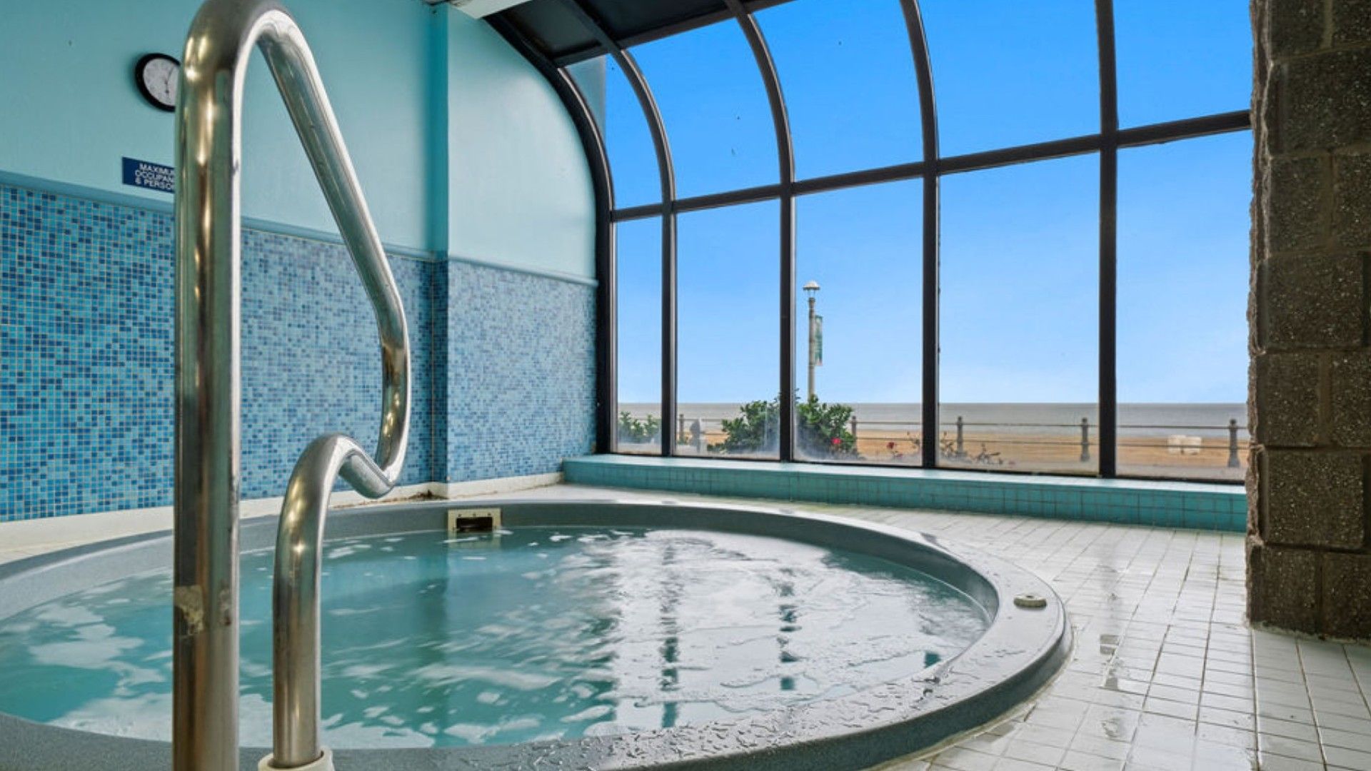 A hot tub with a metal ladder, a tiled wall, and a large window overlooking a beach.