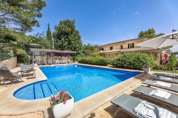 Una piscina amb gandules i una pèrgola, envoltada de vegetació i un edifici.