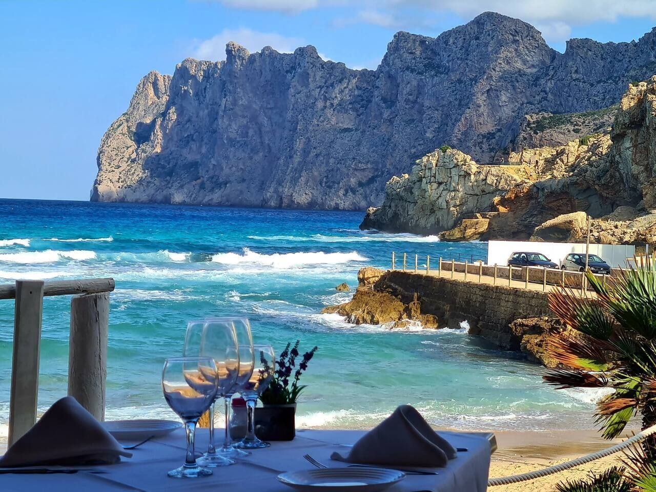 A table set with wine glasses and a small plant, overlooking a rocky coastline with waves and a mountain in the background.