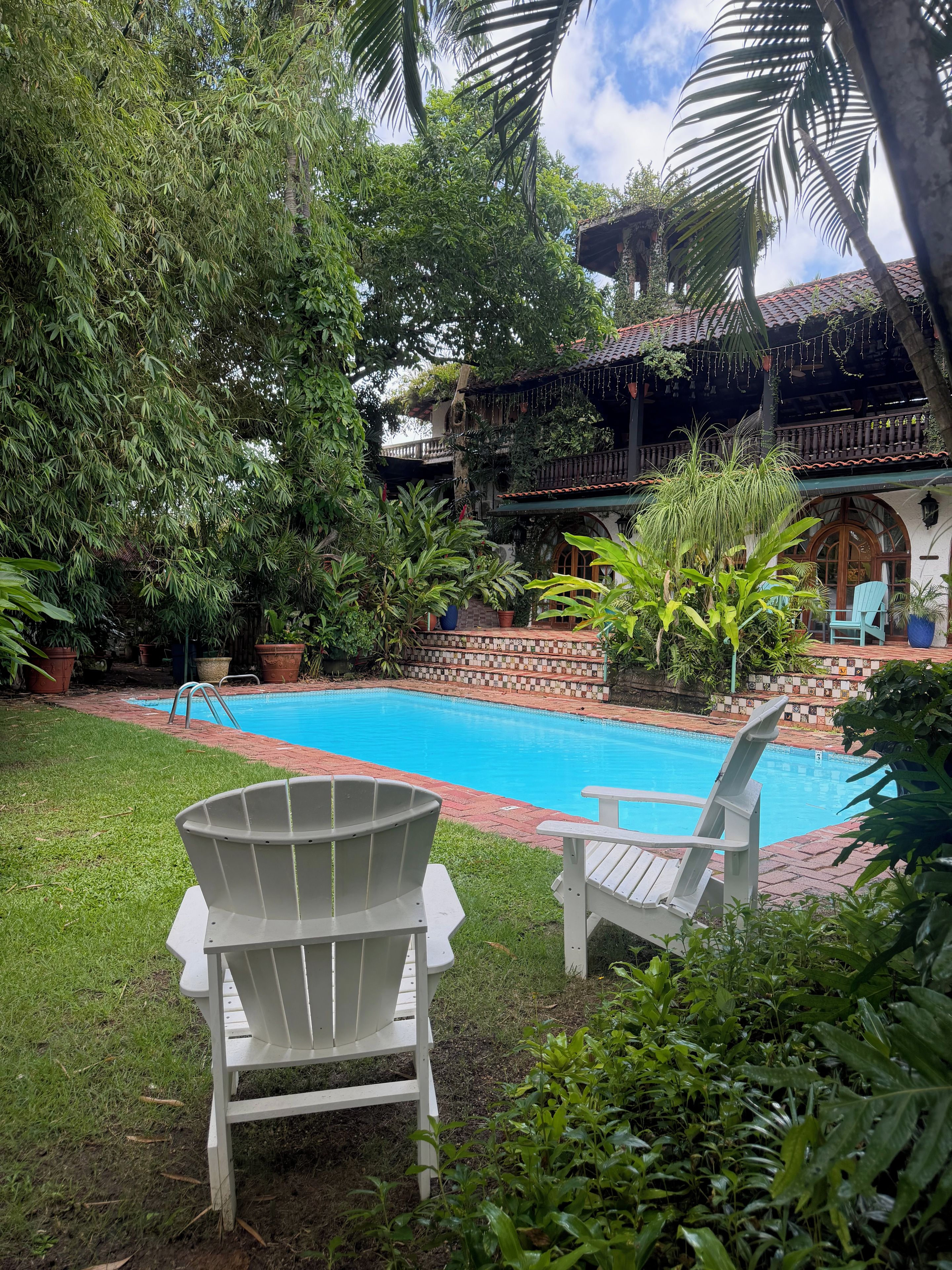 Una piscina con dos sillas Adirondack, rodeada de abundante follaje y un edificio con techo de tejas.