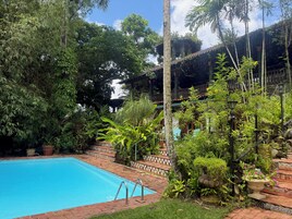 Una piscina rodeada de abundante vegetación y un edificio tradicional con techo de tejas.
