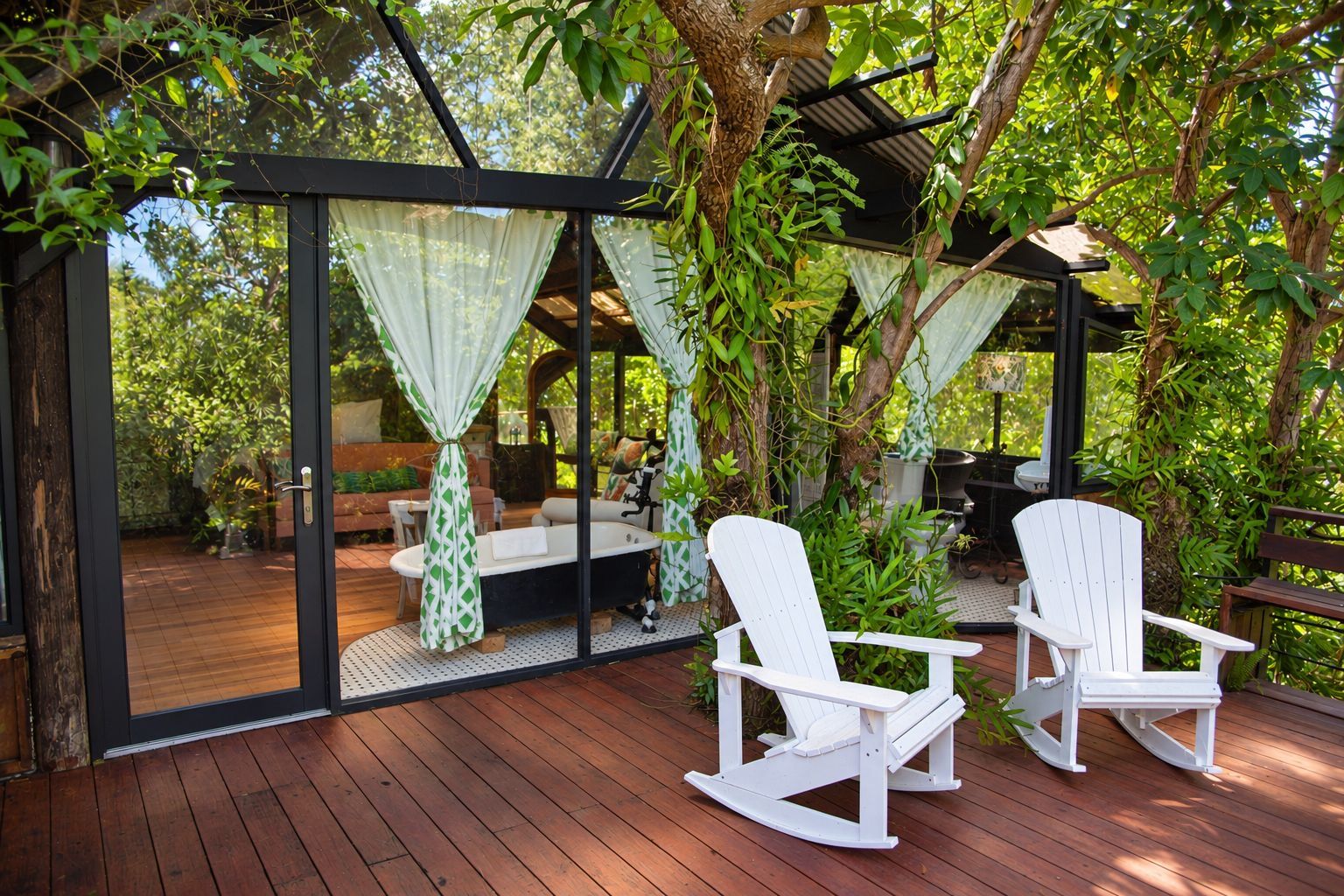 Una terraza de madera con cerramiento de vidrio, mecedoras blancas y un área de descanso con cojines.