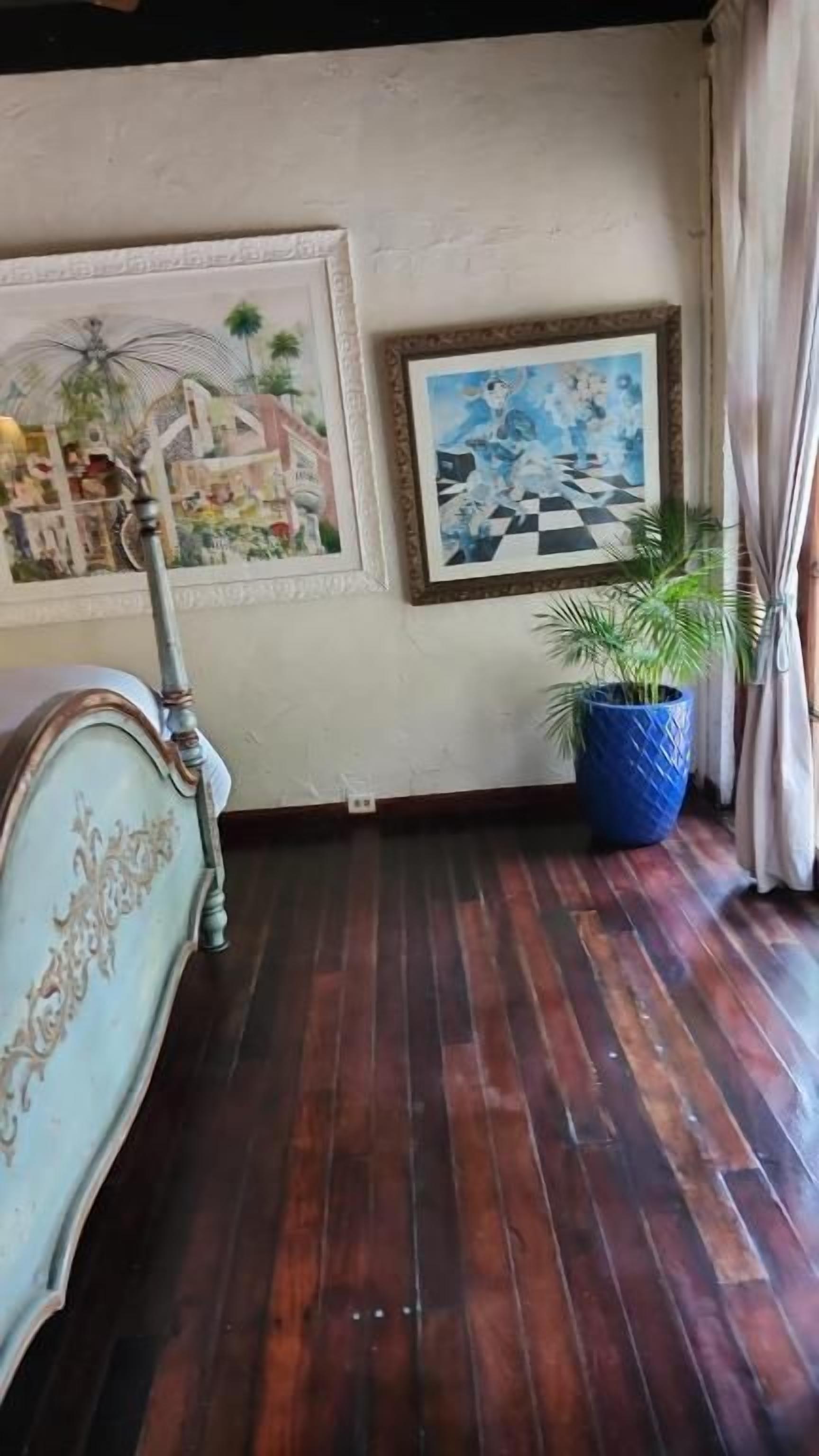 A room with a wooden floor, a bed with an ornate headboard, and two framed artworks on the wall.
