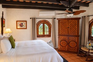 Habitación de hotel con cama, ventilador de techo, ventana de diseño arqueado y aparador de madera con diseños intrincados.