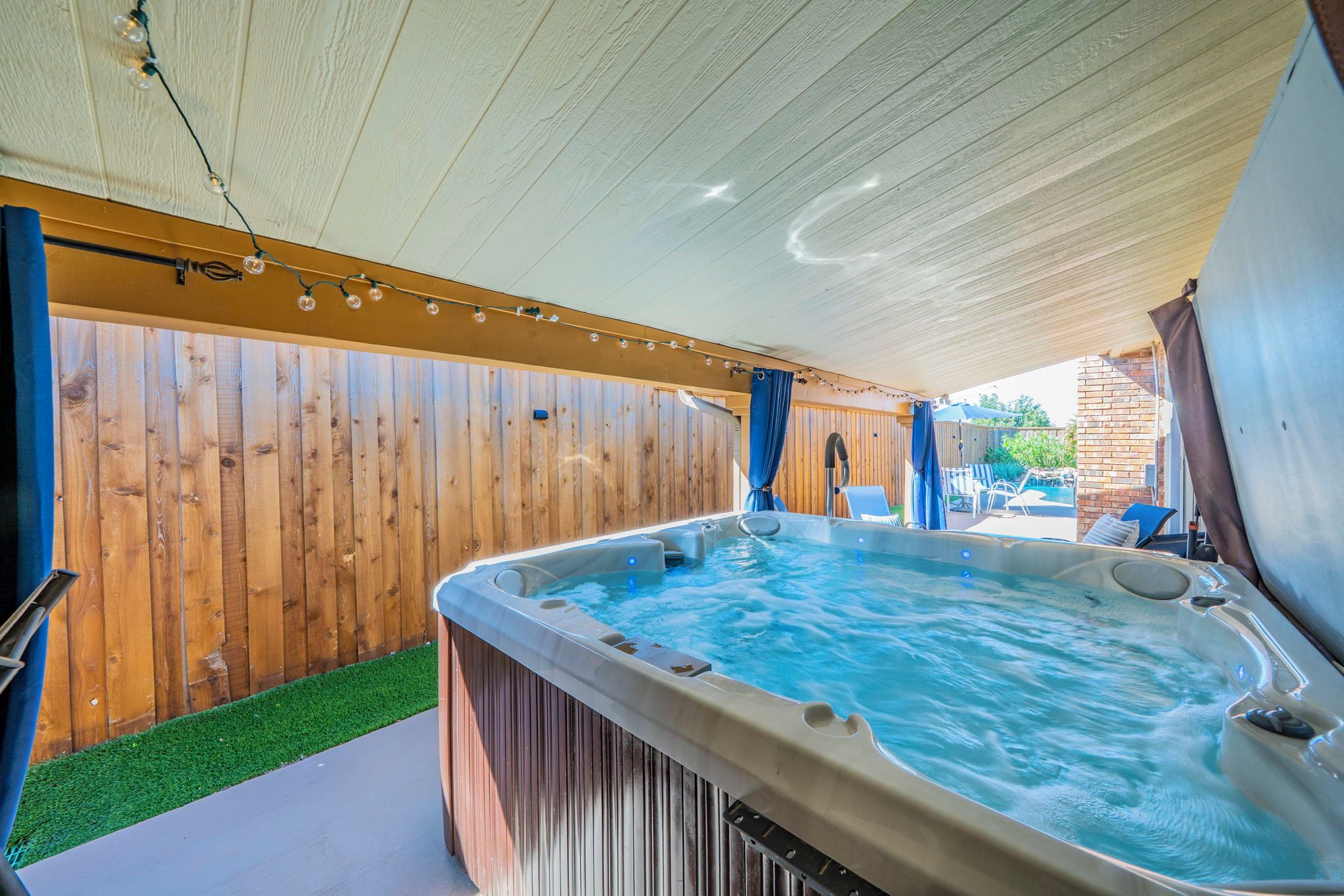 A hot tub under a covered area with a wooden fence and a grassy border.