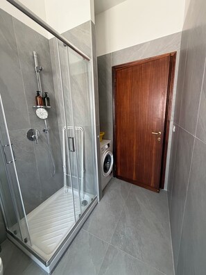A modern bathroom with a glass-enclosed shower, a wooden door, and a washing machine. - New Listing (Roma)